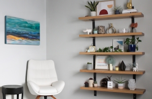 A decorated floating wooden bookshelf hanging on a wall. Next to it is a white leather chair. The floating bookshelves are made of reclaimed barn wood.